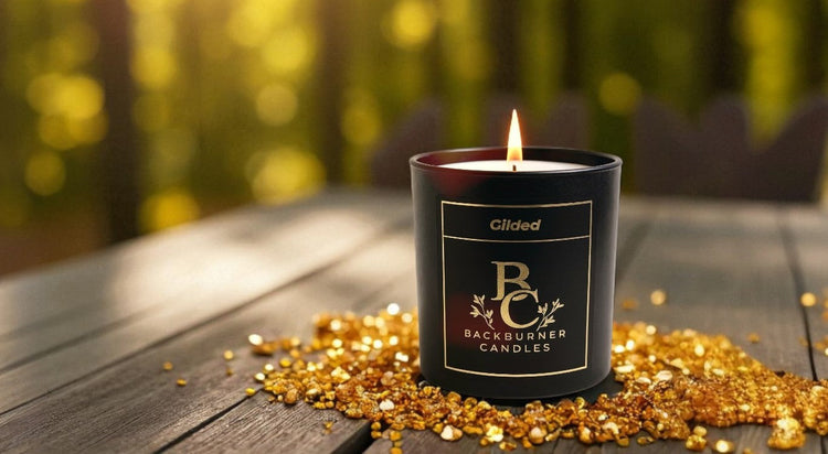 Lit black "Gilded" candle by Backburner Candles on rustic wood outdoors, surrounded by golden glitter, with forest sunlight in background — luxury vegan parasoy wax, inspired by iconic fragrances