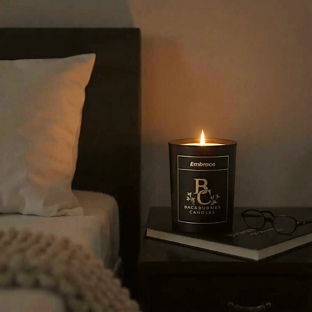 Embrace candle in black glass jar by Backburner Candles on a nightstand next to a bed with a book and glasses