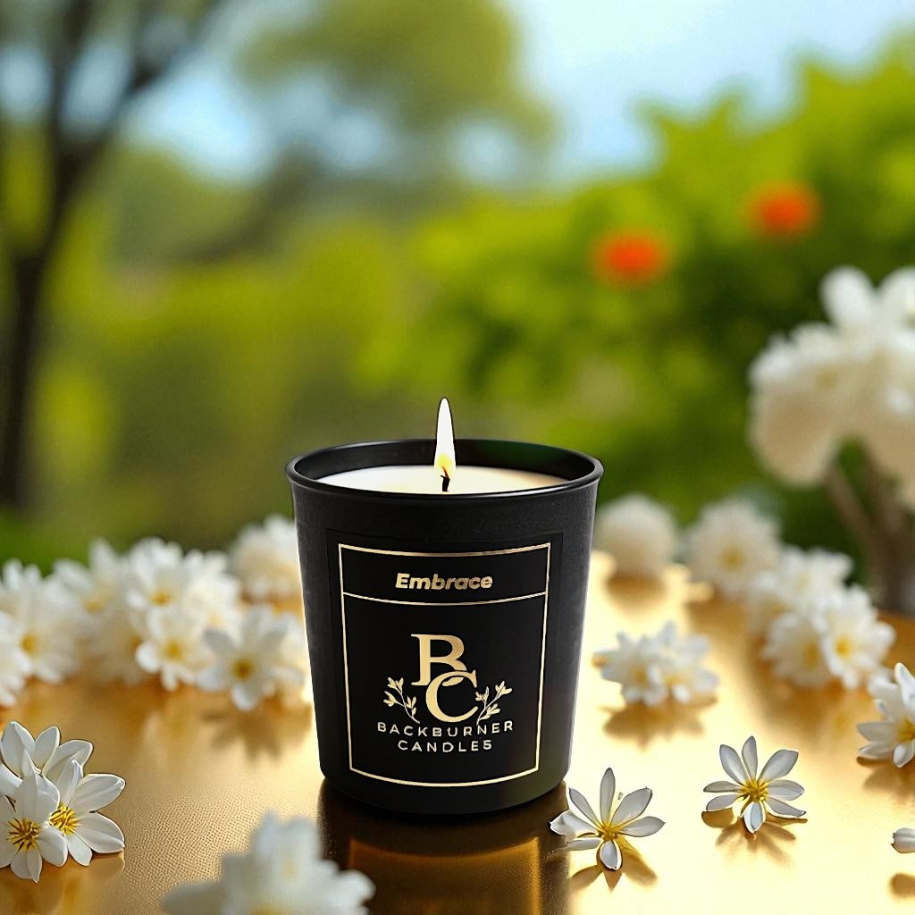 Embrace candle in black glass jar by Backburner Candles surrounded by white flowers outdoors.