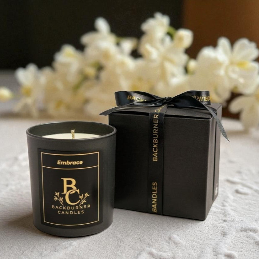 Embrace candle in black glass jar by Backburner Candles and matching packaging box on a textured surface with blurred flowers in the background.