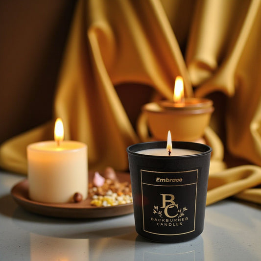 Embrance candle in black glass jar by Backburner Candles on a reflective surface with gold fabric in the background.