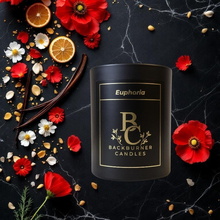 Black candle labeled 'Backburner Candles' surrounded by flowers and fruits on a dark surface