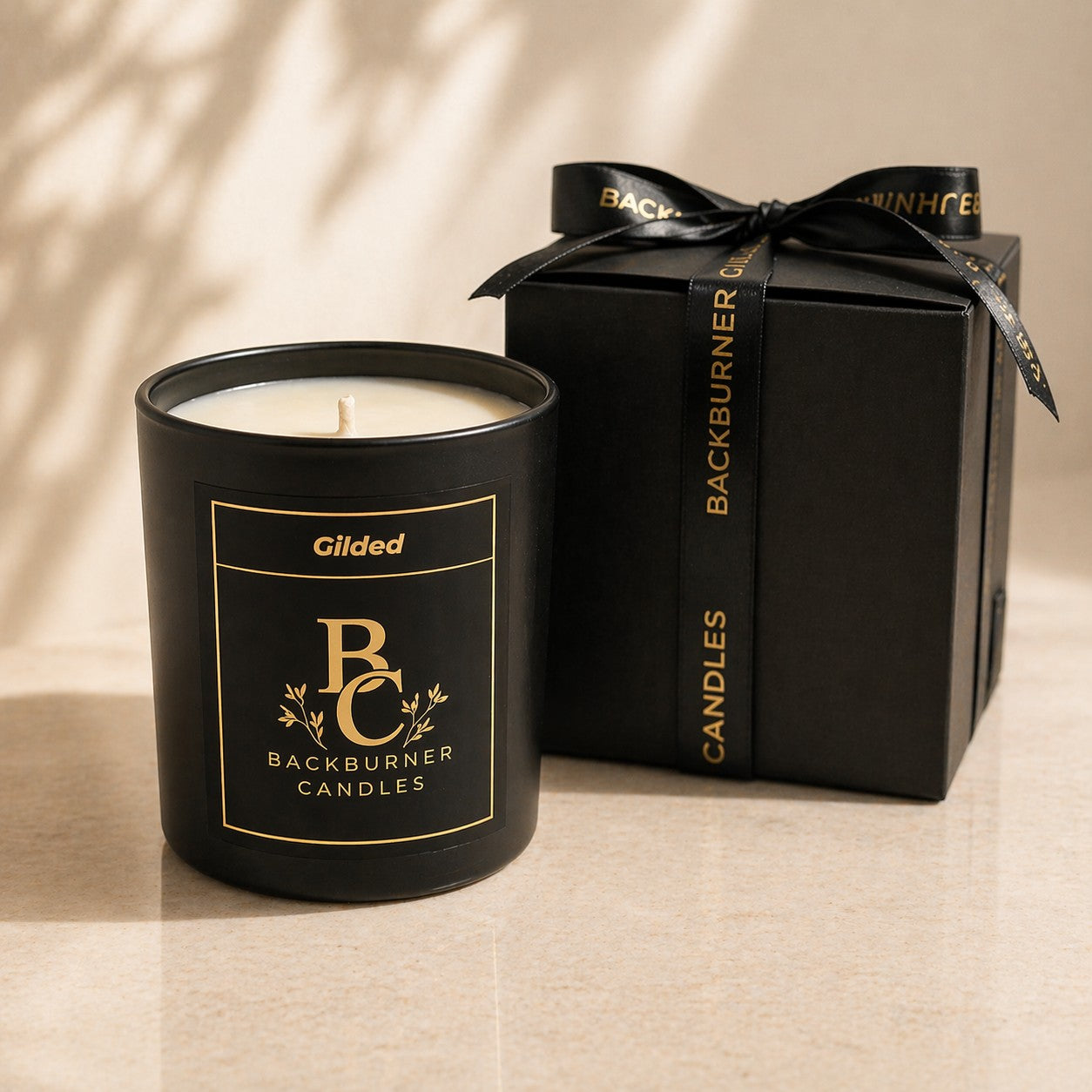 Black candle with gold text next to a matching black gift box on a beige surface