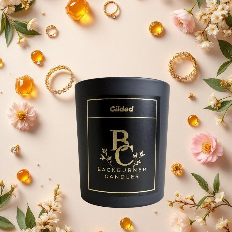 Gilded candle in black glass jar by Backburner Candles surrounded by flowers and jewelry on a light pink background