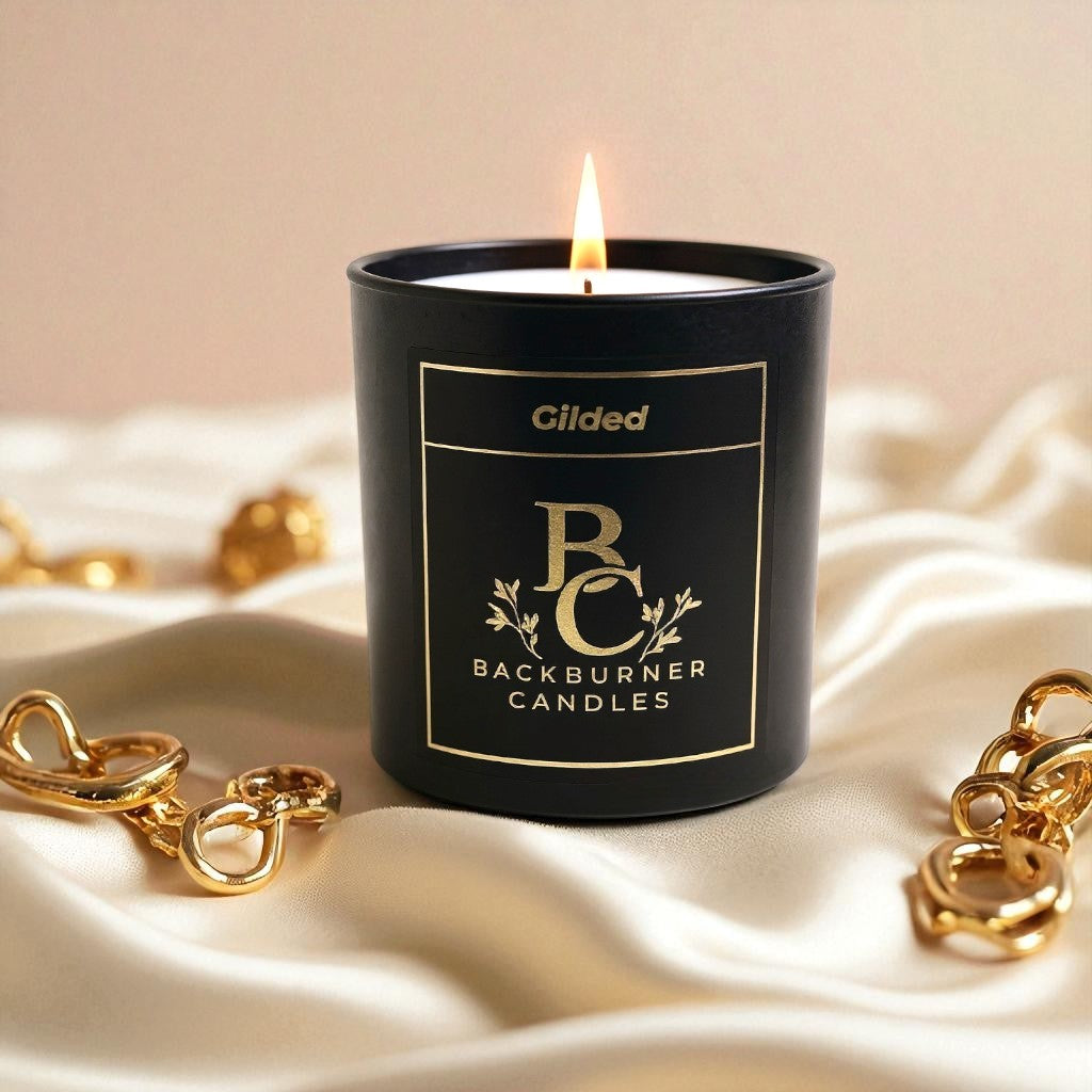 Gilded candle in black glass jar by Backburner Candleson a beige fabric background with gold items