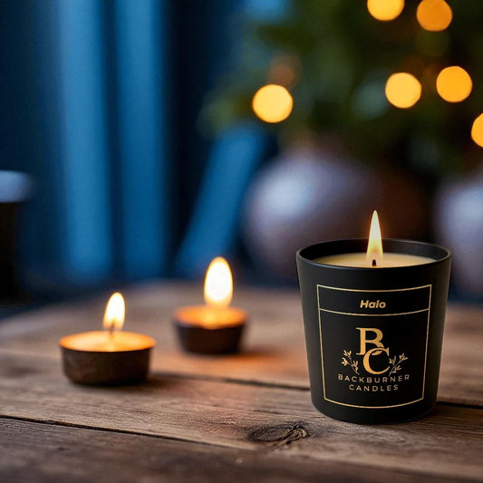 Three candles, including Halo by Backburner Candles in a black glass container, on a wooden surface with a blurred festive background.