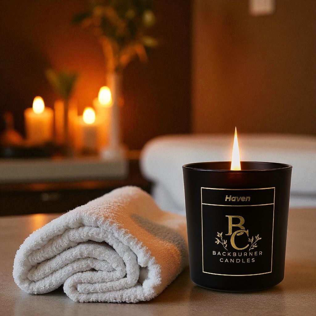Haven candle in black glass jar by Backburner Candles next to a folded towel on a blurred background of candles and plants.