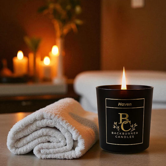 Haven candle in black glass jar by Backburner Candles next to a folded towel on a blurred background of candles and plants.