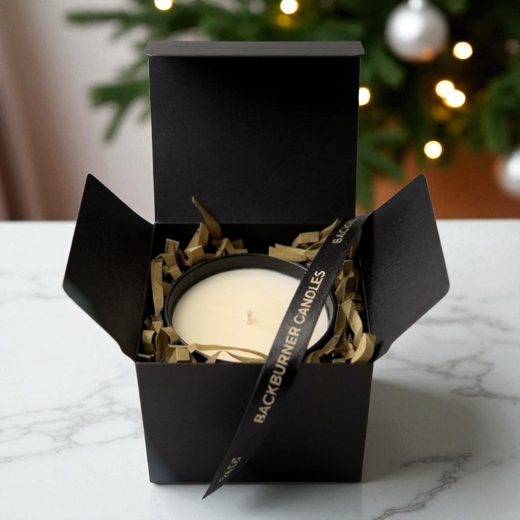 White candle in black gift box with gold ribbon and paper shreds, styled for elegant gifting by Backburner Candles.