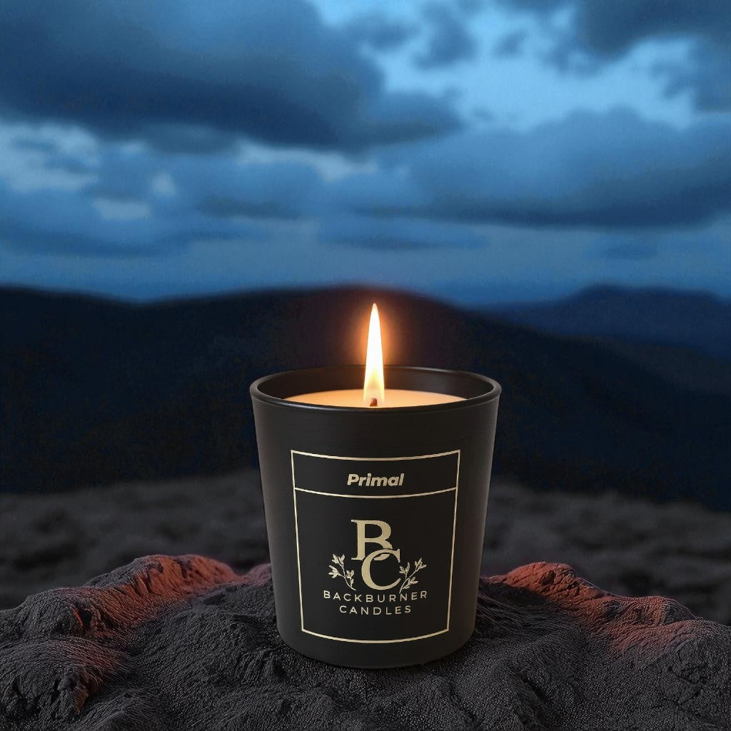 Primal candle in a black glass jar against a dark mountainous landscape.