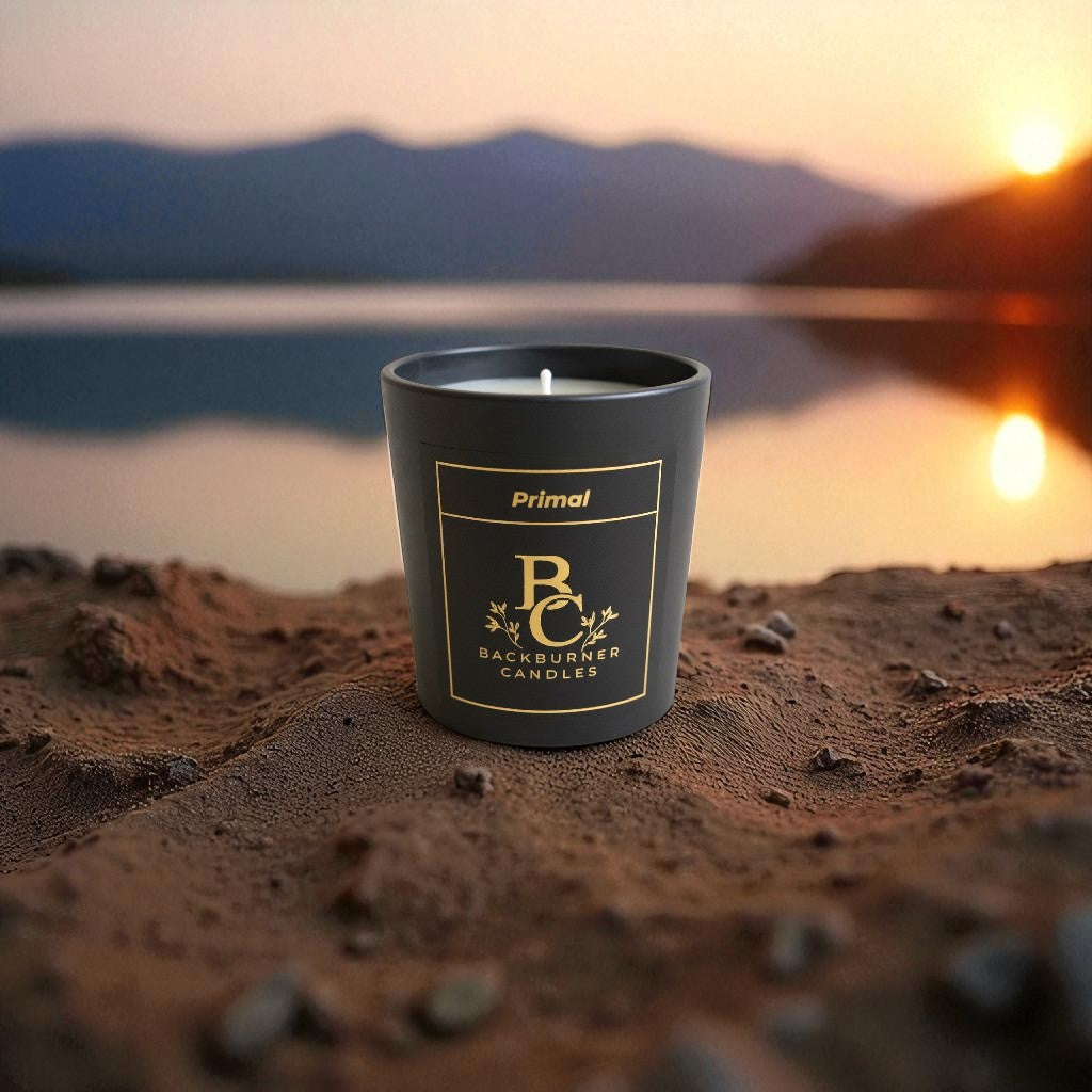 Primal candle by Backburner Candles in a black container on sand with sunset or sunrise in the background.