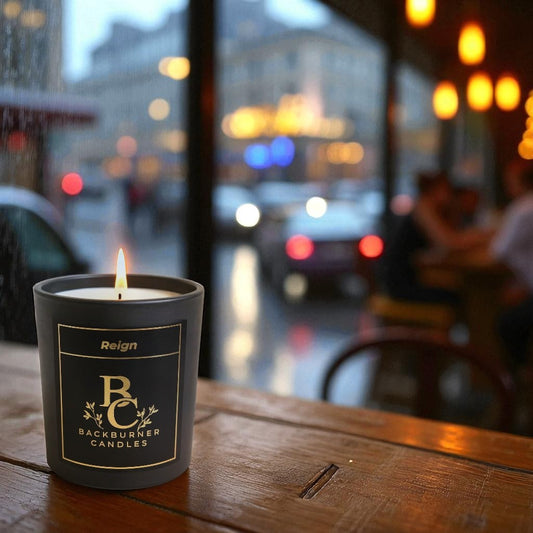 Backburner Candles Reign candle in a black container on a wooden table, blurred city street in the background.