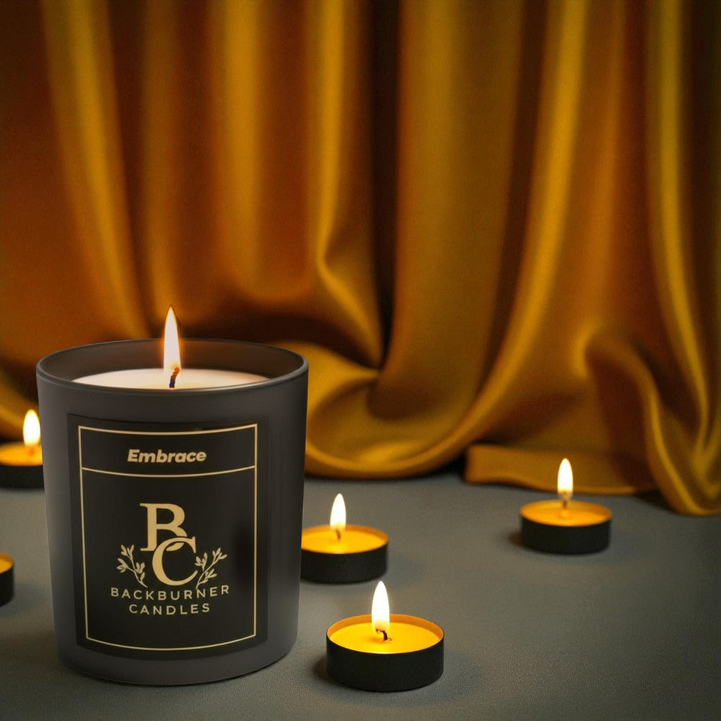Embrace candle in black glass jar by Backburner Candles on a reflective surface with gold fabric in the background.