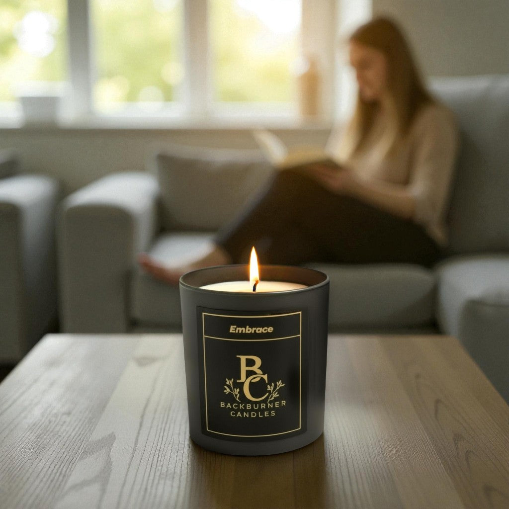 Lit candle in a black container with 'Backburner Candles' branding on a wooden table, with a blurred person reading in the background.