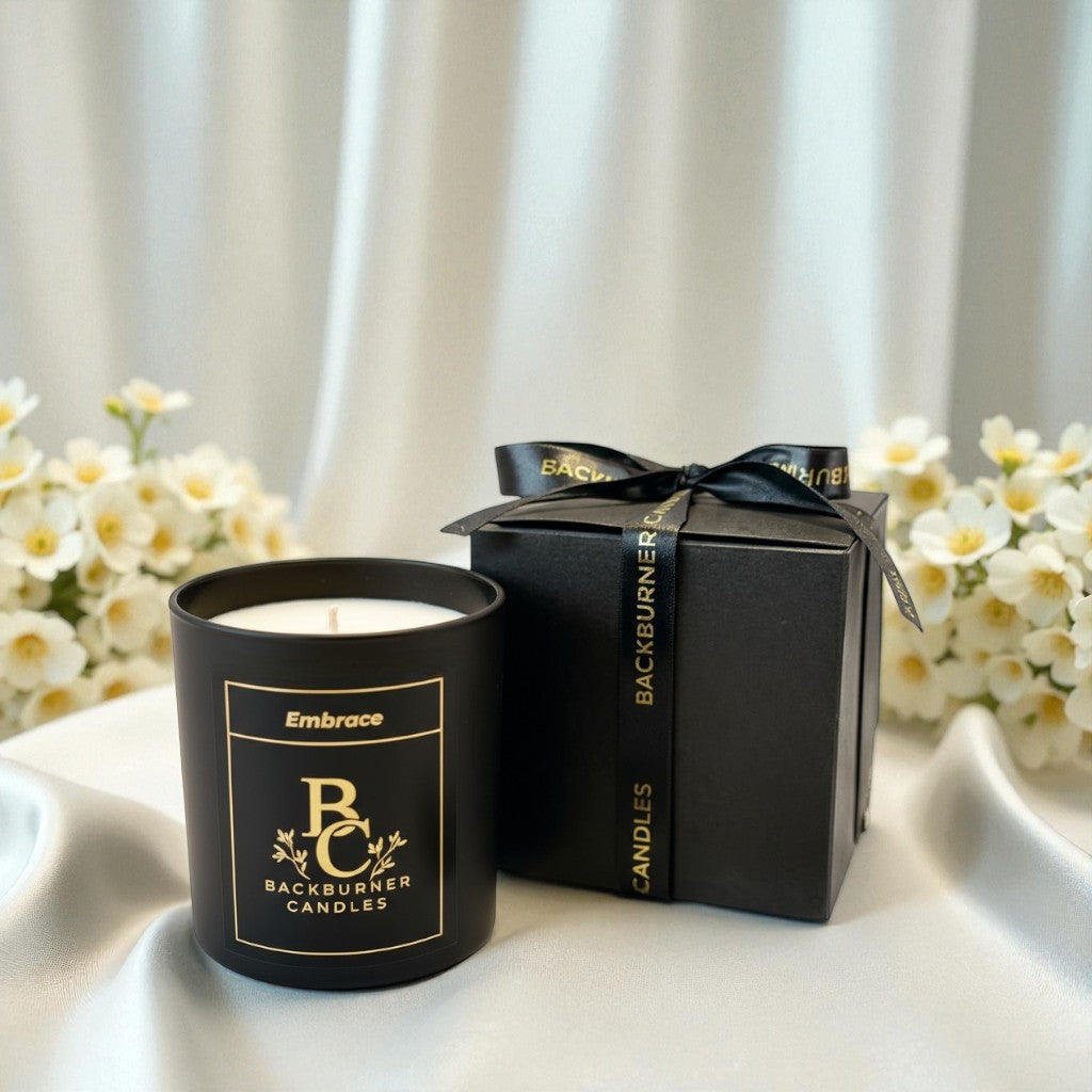 Black candle with gold text and a matching black gift box on a white surface with flowers in the background