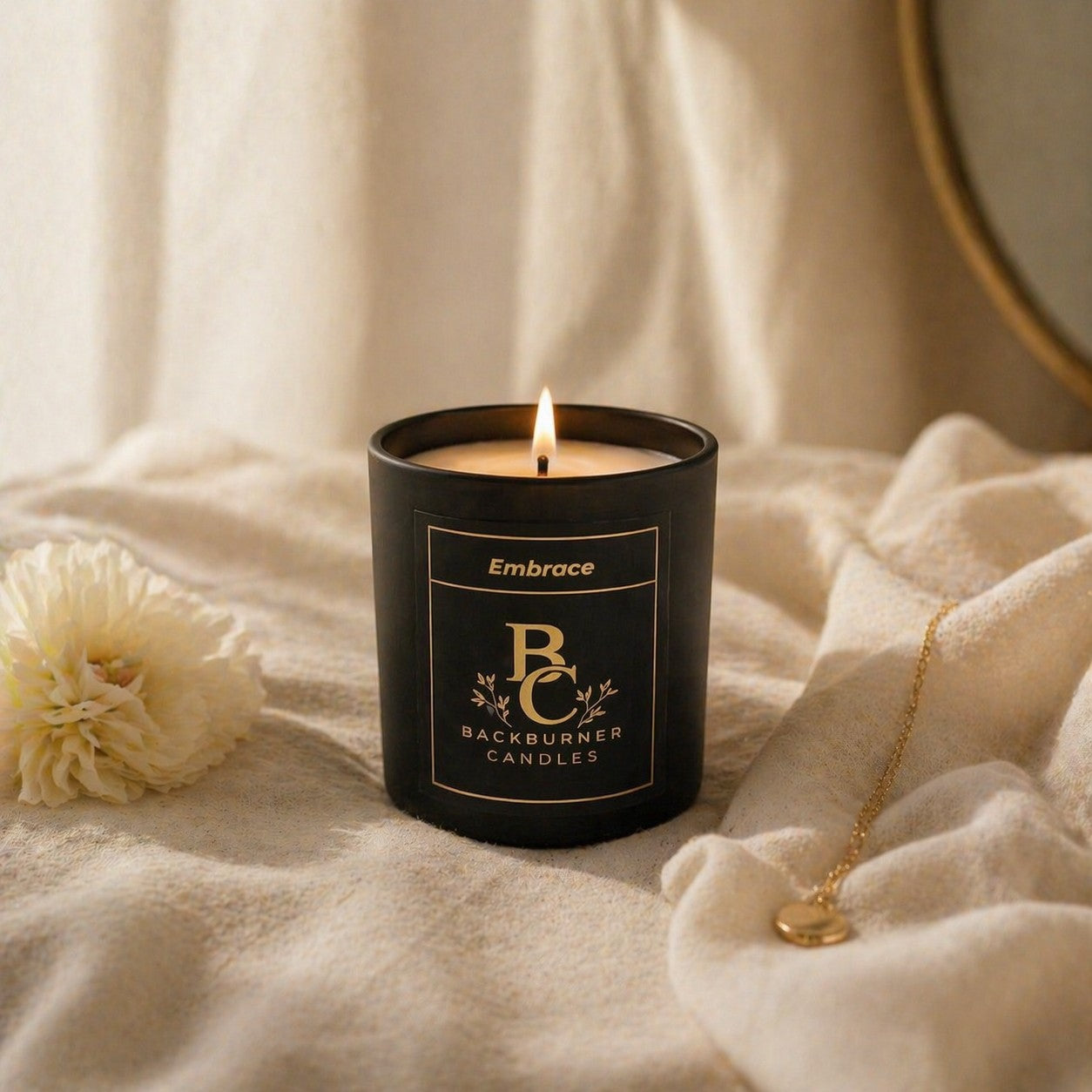 Candle in a black container with 'Embrace' branding on a soft fabric background