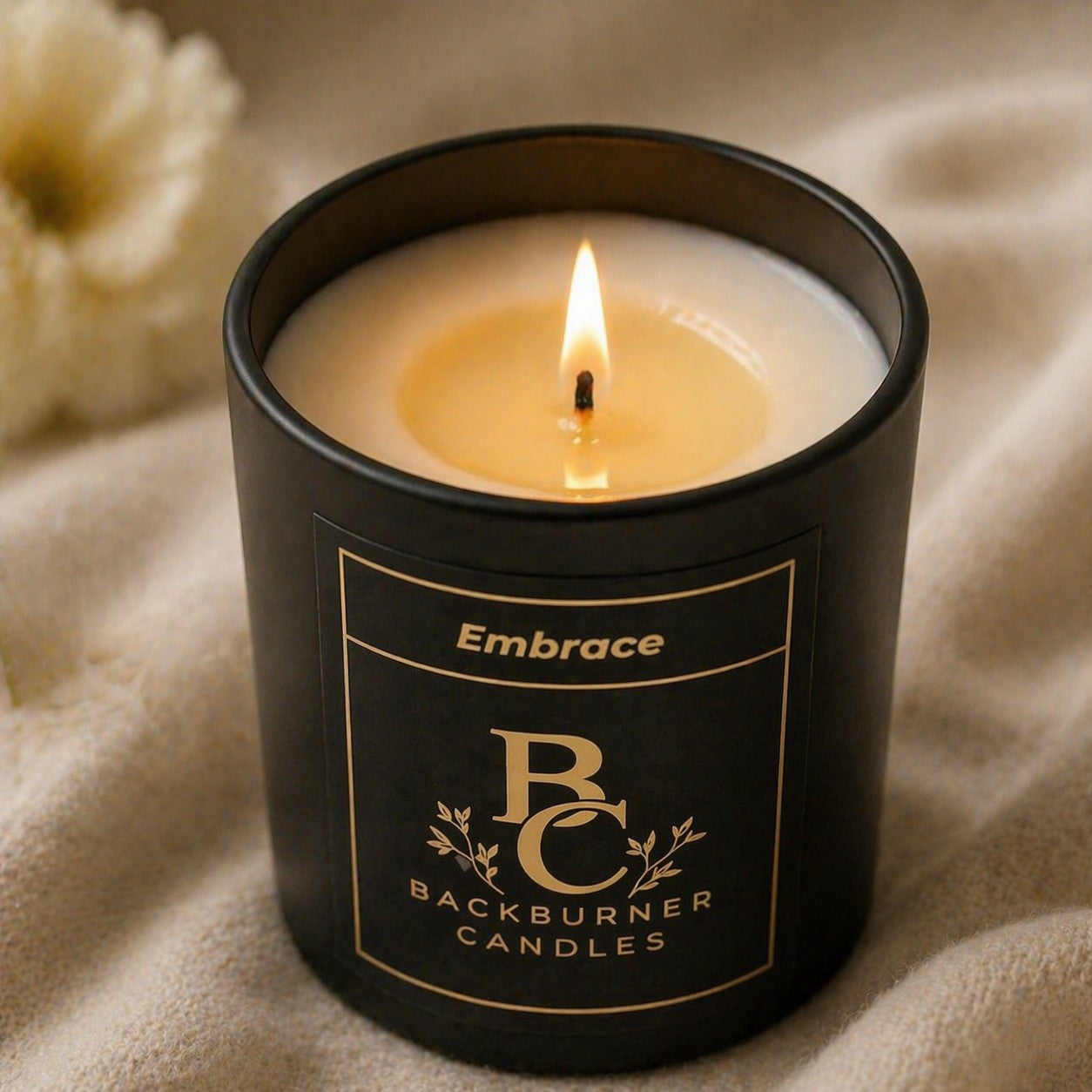 Black candle in a container with 'Embrace Backburner Candles' label on a beige fabric background