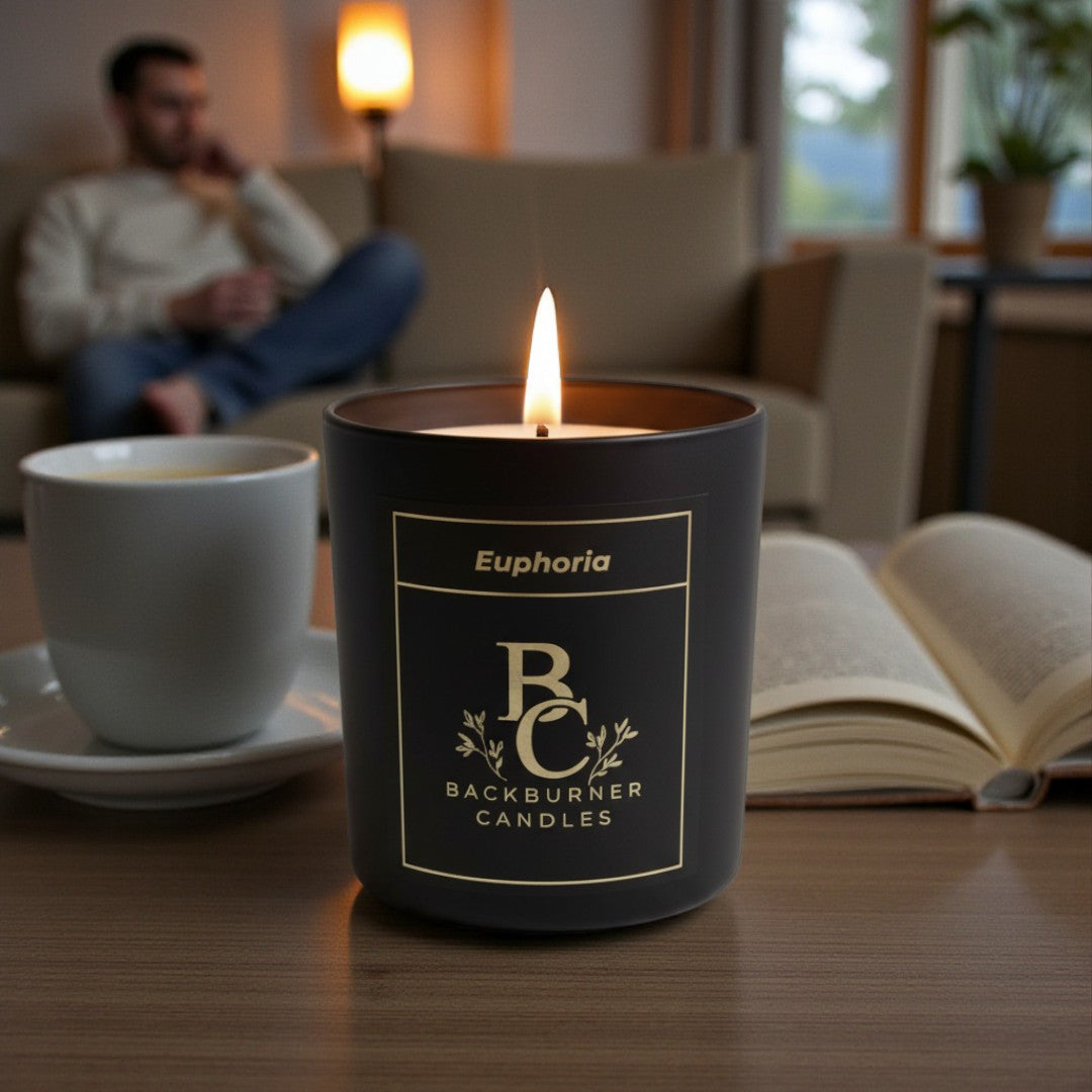 Candle with 'Backburner Candles' branding on a table with a cup and book in a cozy room.