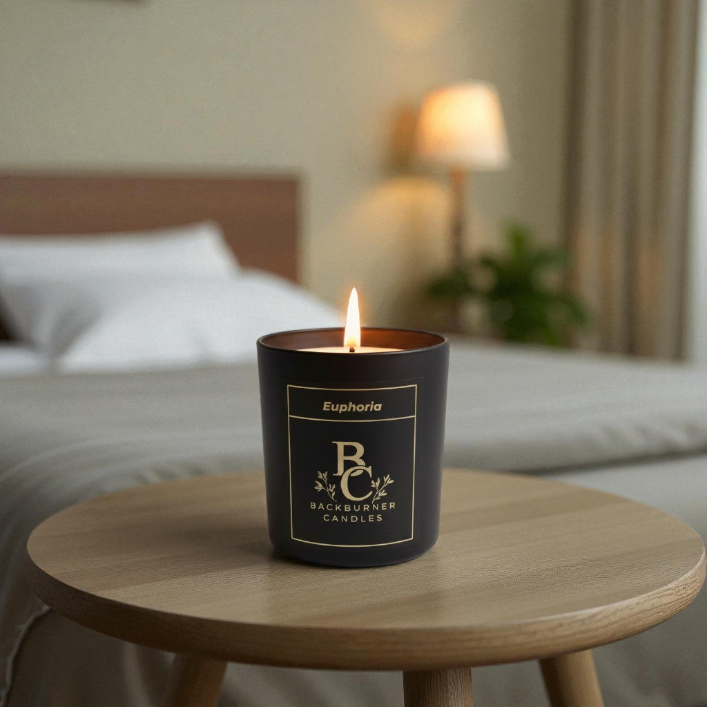 Candle in a black container labeled 'Backburner Candles' on a wooden table with a blurred bedroom background.