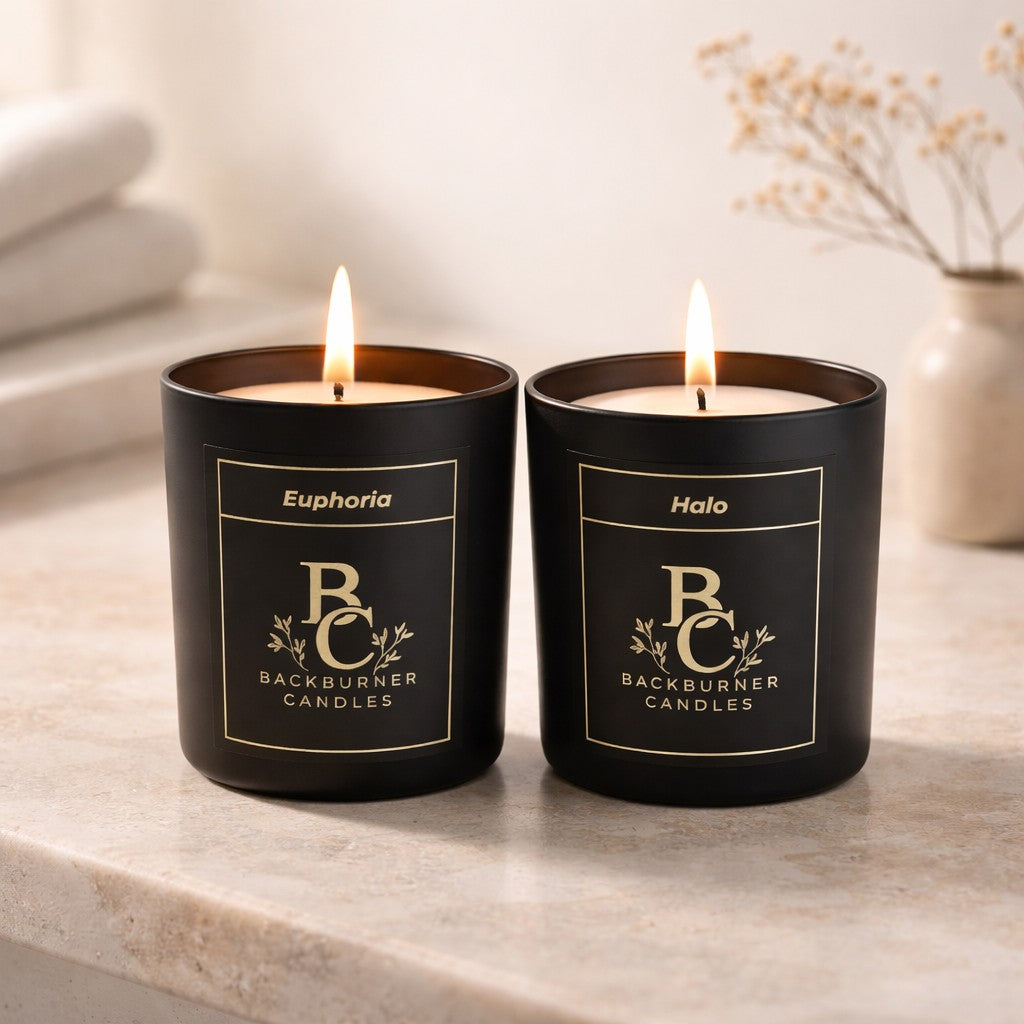 Two lit candles in black containers with visible labels on a marble. surface. The February Comfort Pair.