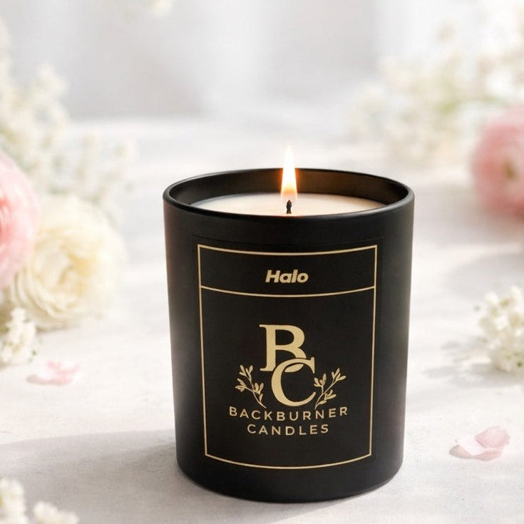 Black candle with 'Backburner Candles' branding on a white surface with flowers in the background