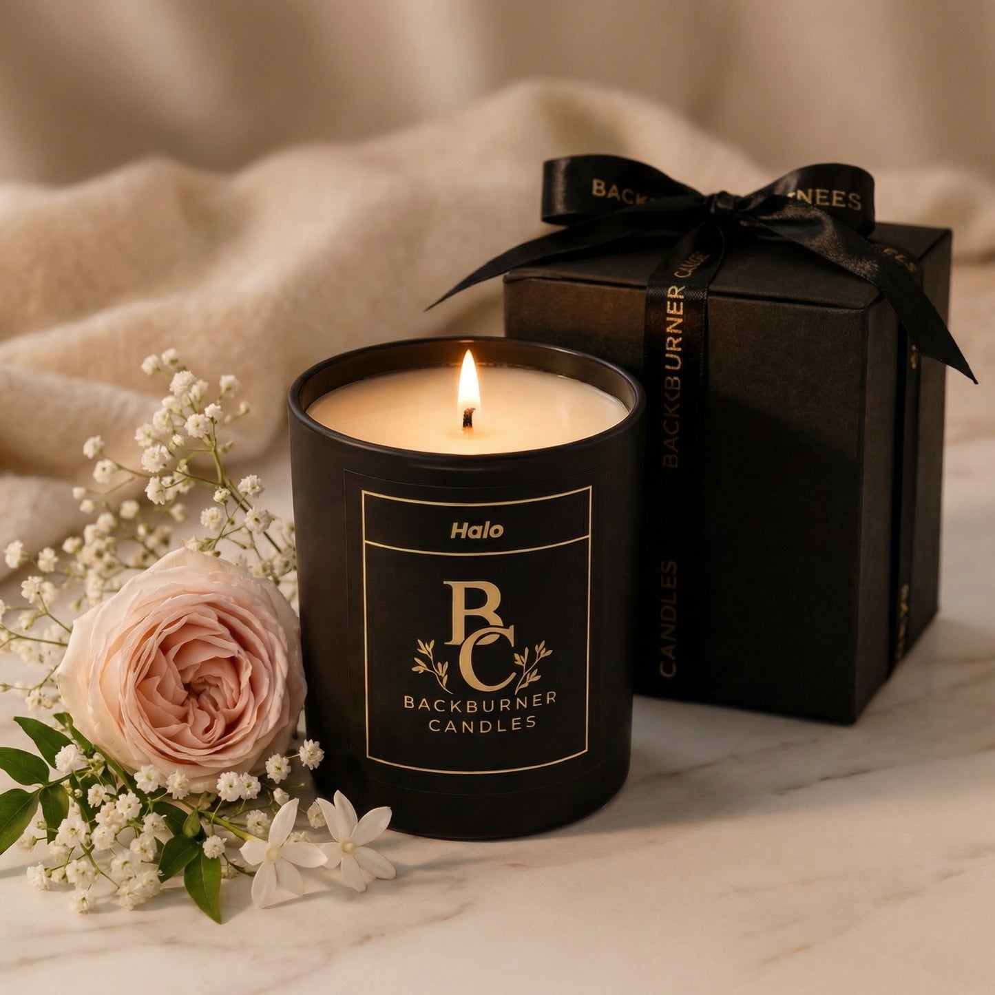 Black candle with 'Backburner Candles' branding, surrounded by flowers and a gift box.