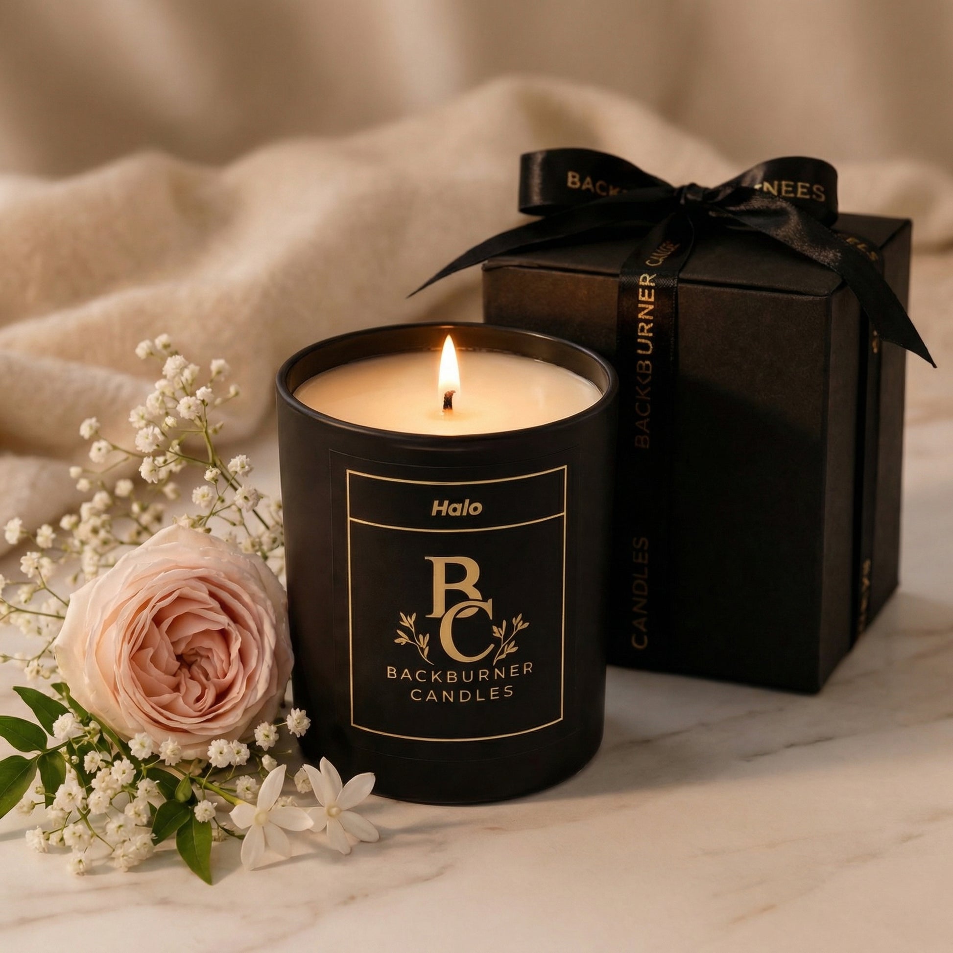 Black candle with 'Backburner Candles' branding, surrounded by flowers and a gift box.