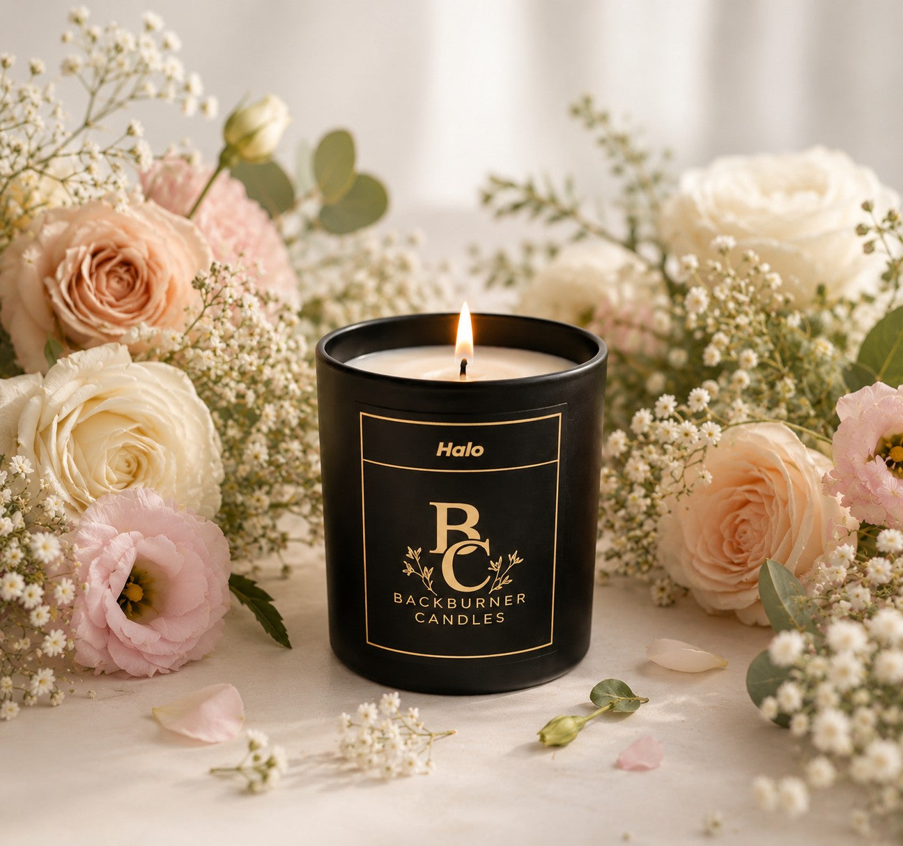 Black candle with 'Backburner Candles' branding surrounded by flowers on a light surface.