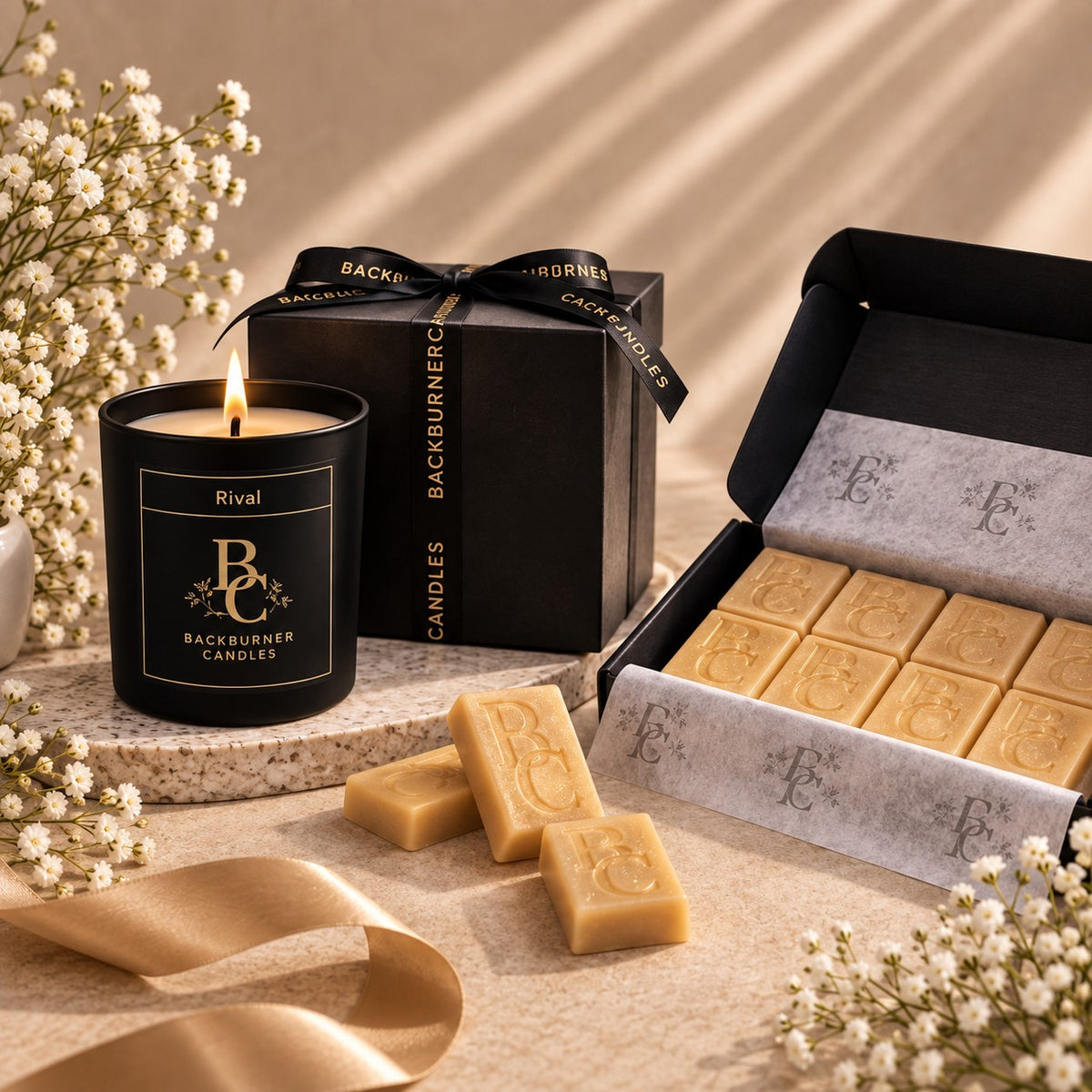 A luxury candle gift set from Rival Backburner Candles, featuring a black glass jar candle with a lit flame, a black gift box with gold ribbon, and an open box of natural unbleached beeswax bars, styled with dried baby's breath flowers on a beige stone surface.