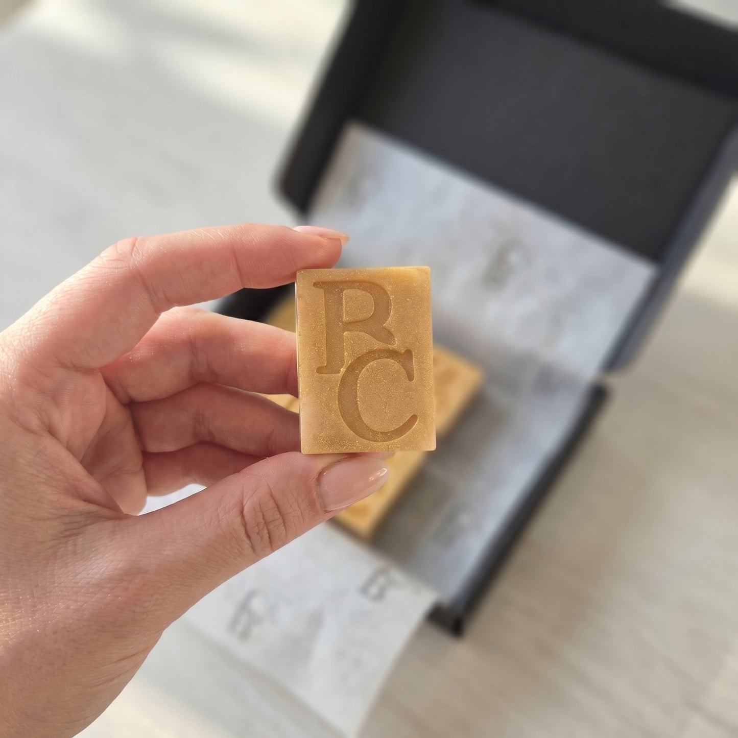 Hand holding a rectangular wax melt with 'BC' branding against a blurred background