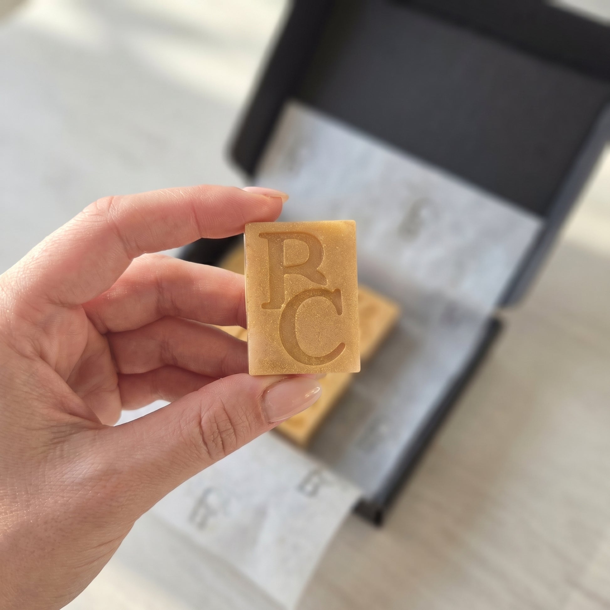 Hand holding a rectangular wax melt with 'BC' branding against a blurred background