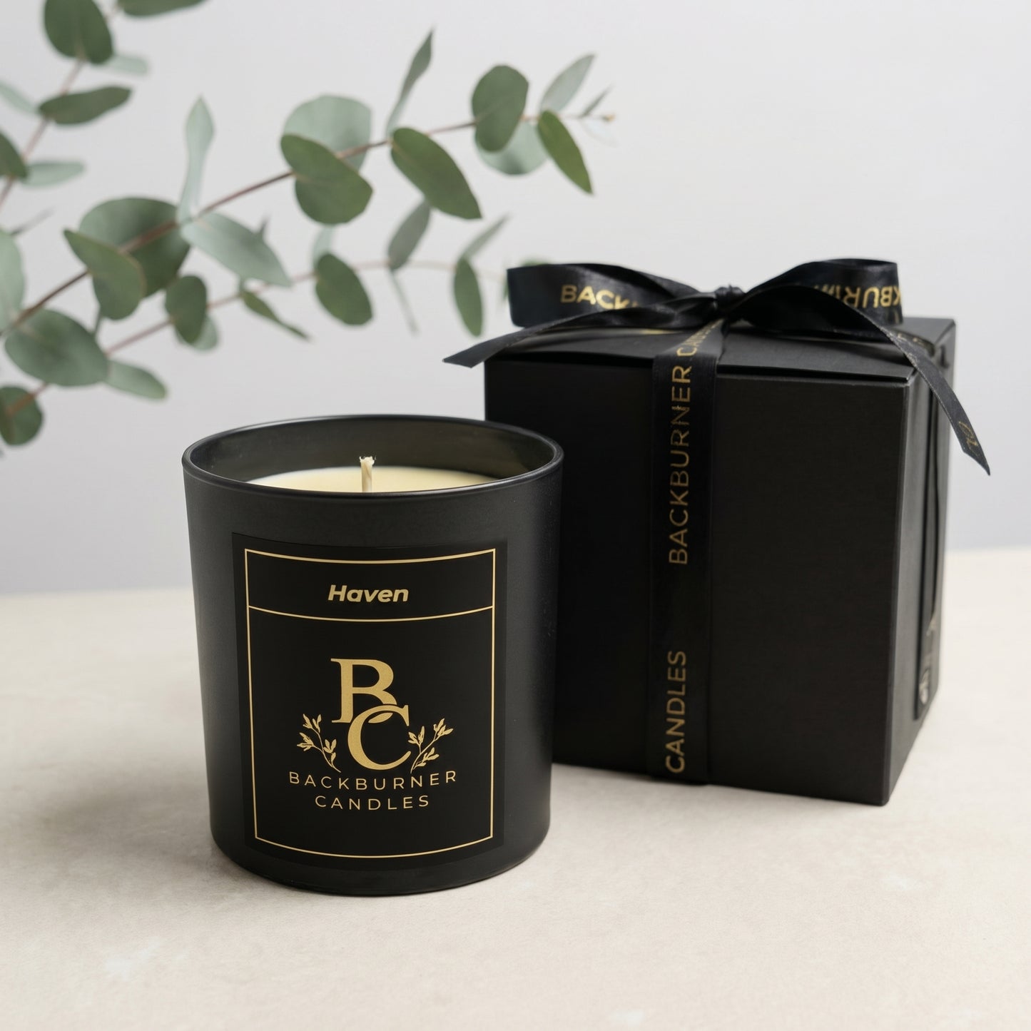 Black candle with 'Backburner Candles' branding next to a black gift box on a light surface.