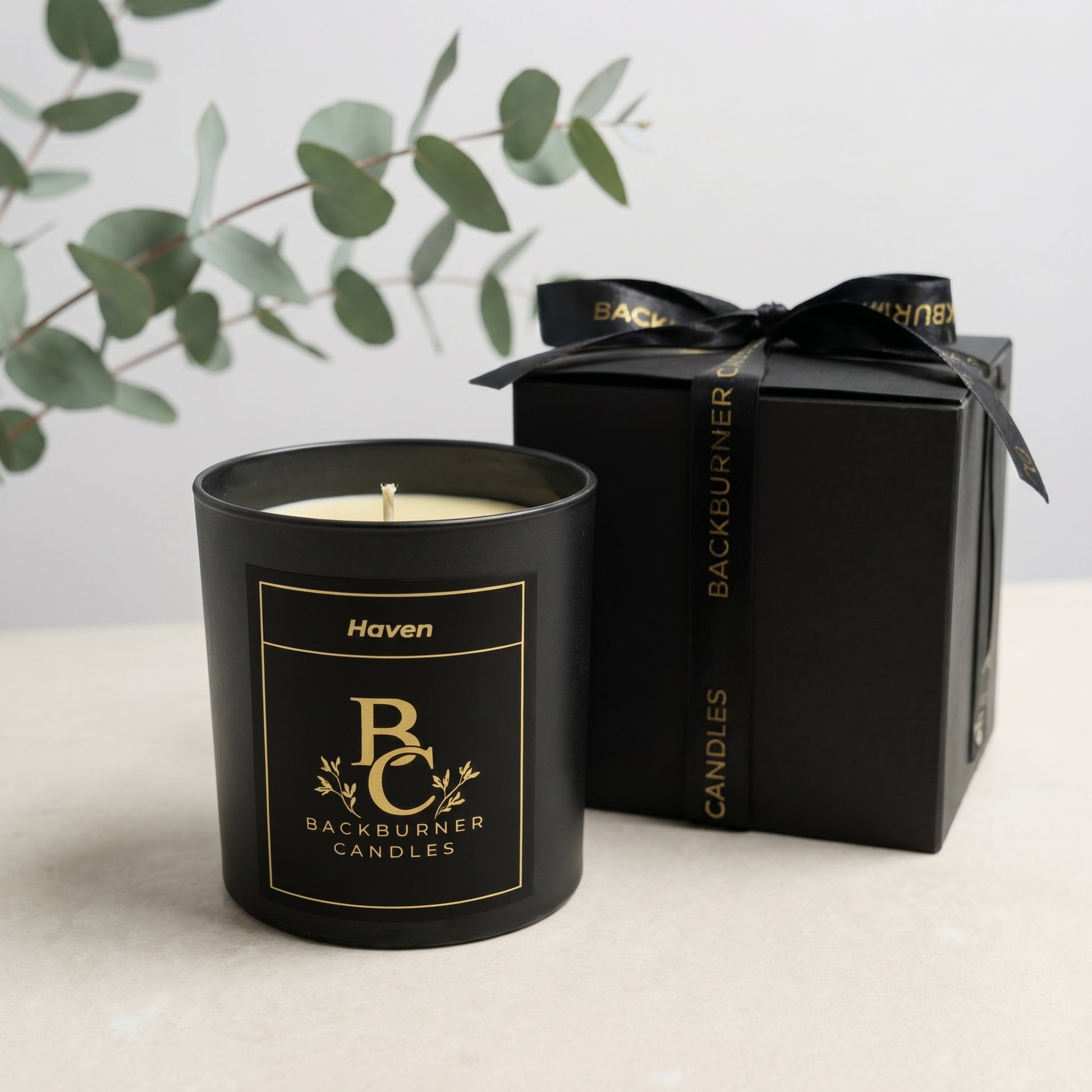 Black candle with 'Backburner Candles' branding next to a black gift box on a light surface.