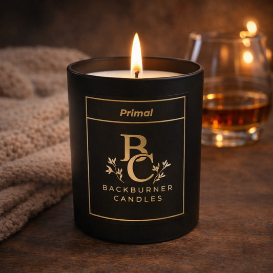 Black candle with 'Backburner Candles' label and 'Primal' branding, lit with a glass of whiskey in the background.