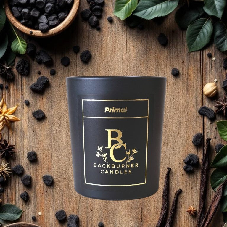 Black candle with 'Primal Backburner Candles' label on a wooden surface with dark fruits and leaves.