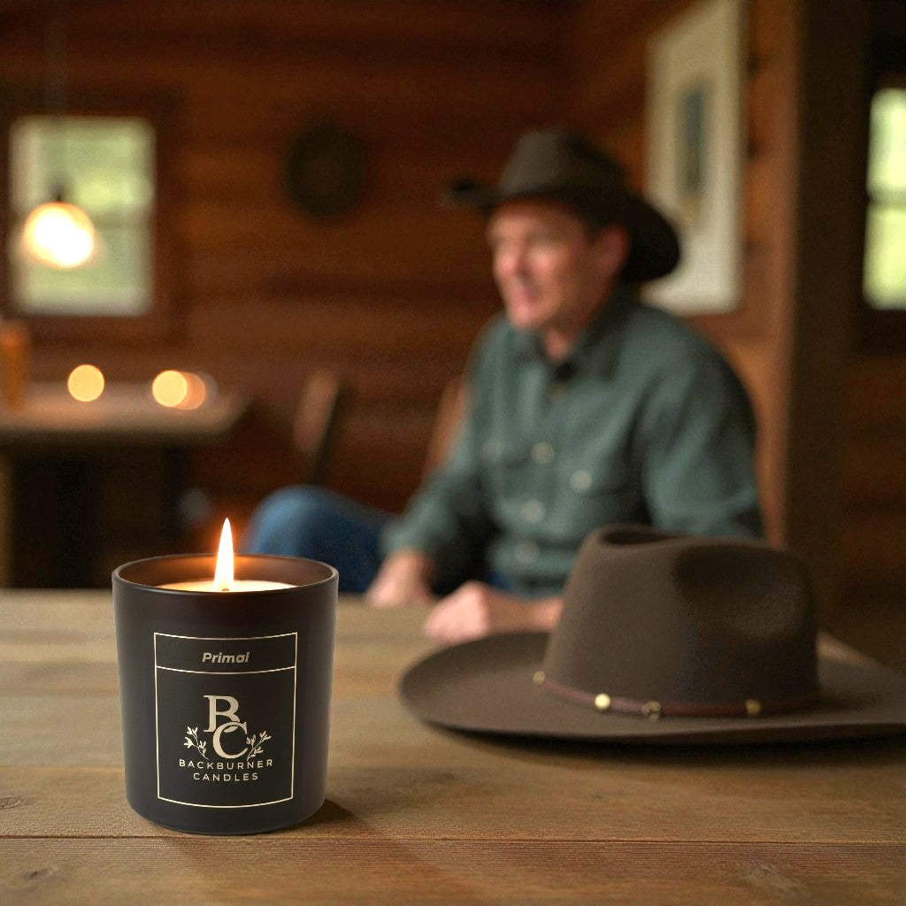Candle with 'BC' branding on a wooden table, man in background wearing a hat.