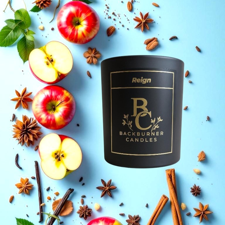 Black candle with 'Reign Backburner Candles' branding surrounded by apples, cinnamon sticks, and star anise on a blue background.