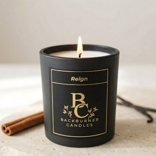 Black candle with 'Reign Backburner Candles' label on a light surface with vanilla beans and cinnamon sticks.