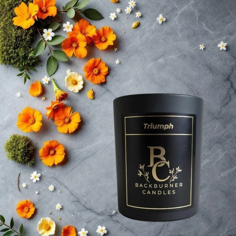 Black candle with 'Backburner Candles' branding surrounded by flowers on a gray surface