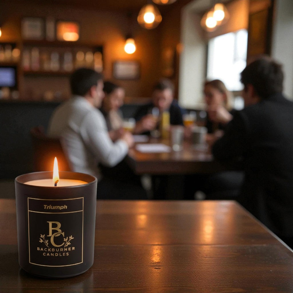 Lit Triumph vegan candle by Backburner Candles on wooden table, styled in cosy bar setting with warm lighting and social ambiance.