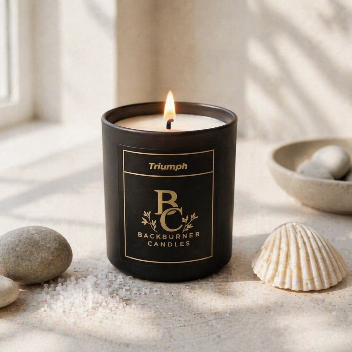 Candle in a black container labeled 'Triumph Backburner Candles' on a light surface with stones and a shell.
