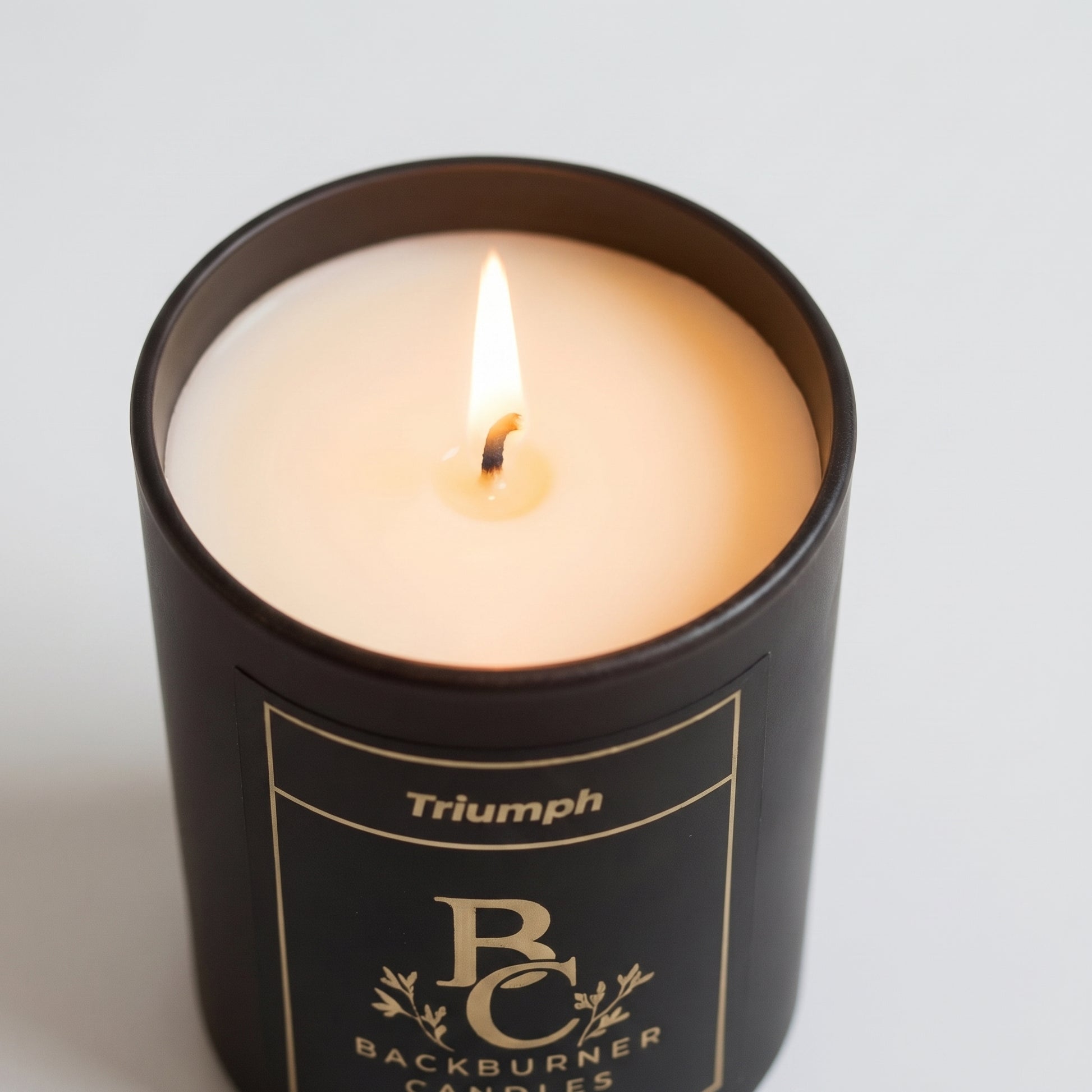 Candle in a black container with 'Triumph' branding on a light gray background