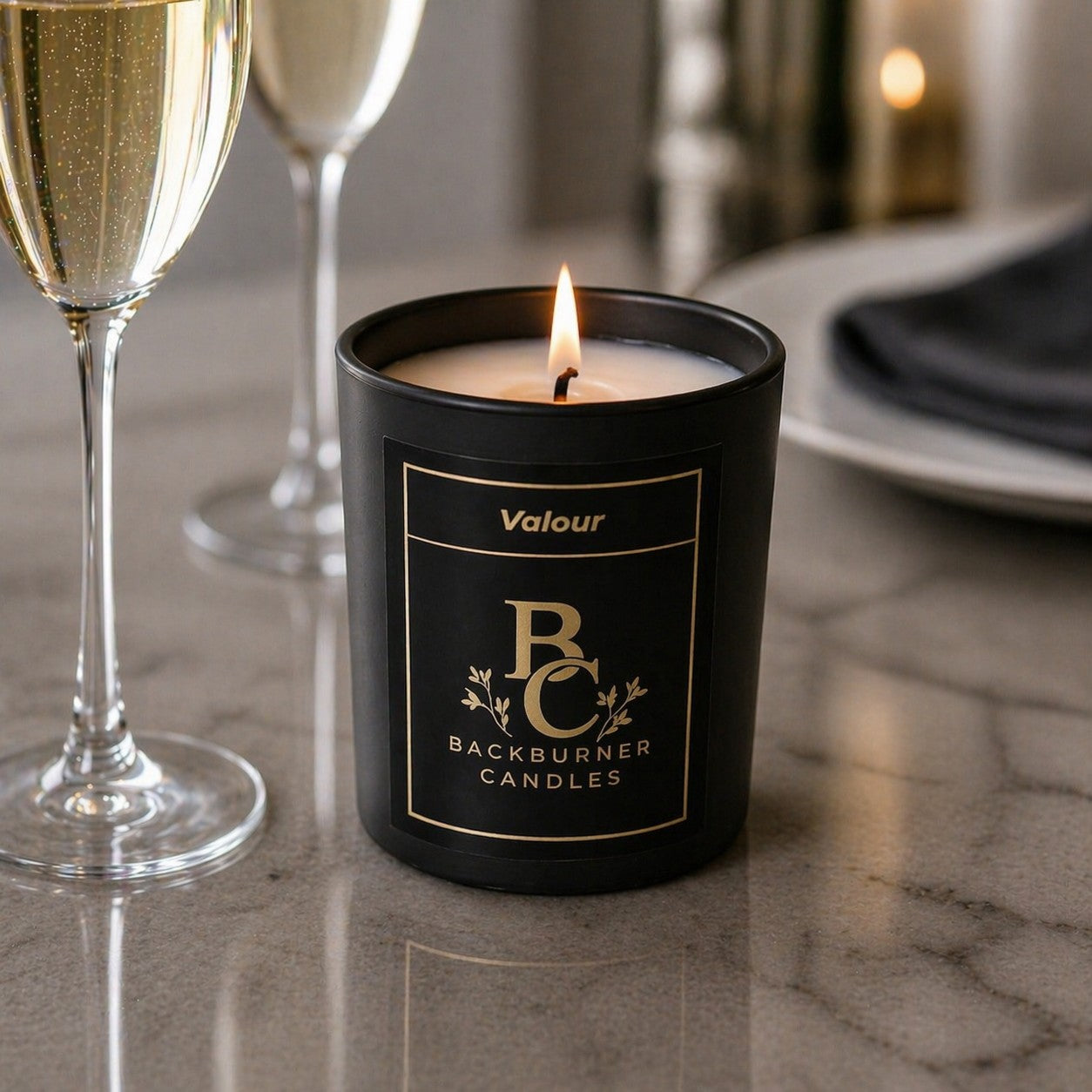 Lit candle in a black container with 'Backburner Candles' branding on a table with champagne glasses.