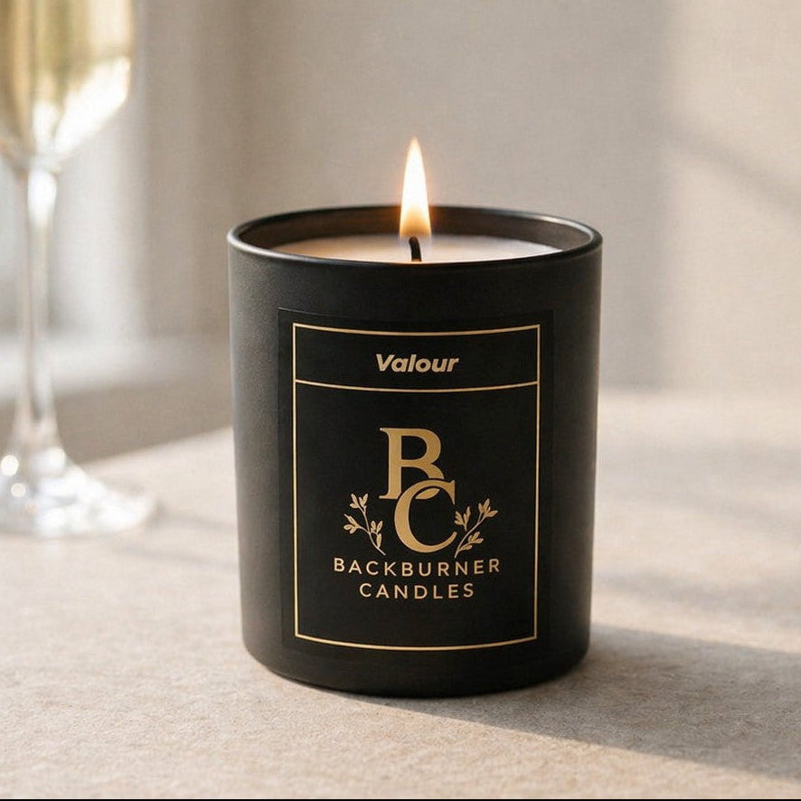 Black candle with 'Backburner Candles' branding on a light surface, next to a glass of champagne.