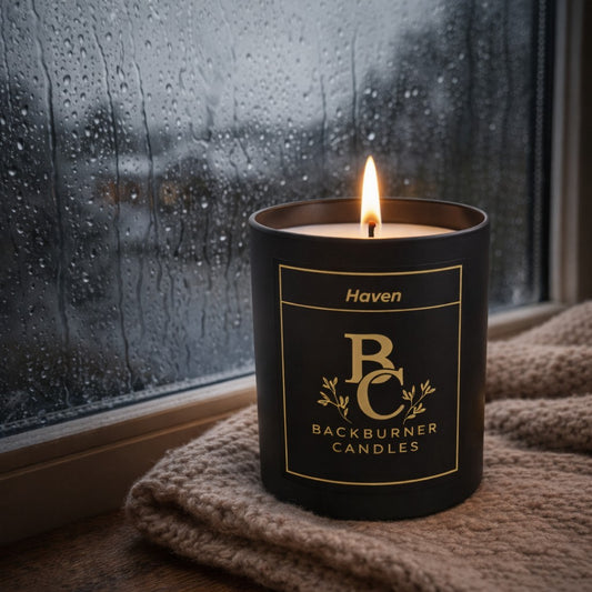 Candle in a black container with 'Backburner Candles' branding, lit in front of a rainy window.
